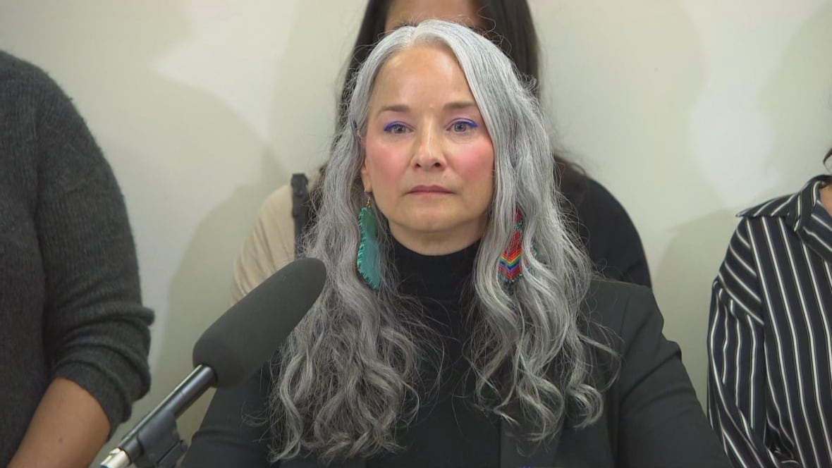 A woman with long grey hair and rainbow beaded earrings stands in front of a microphone at a government announcement.
