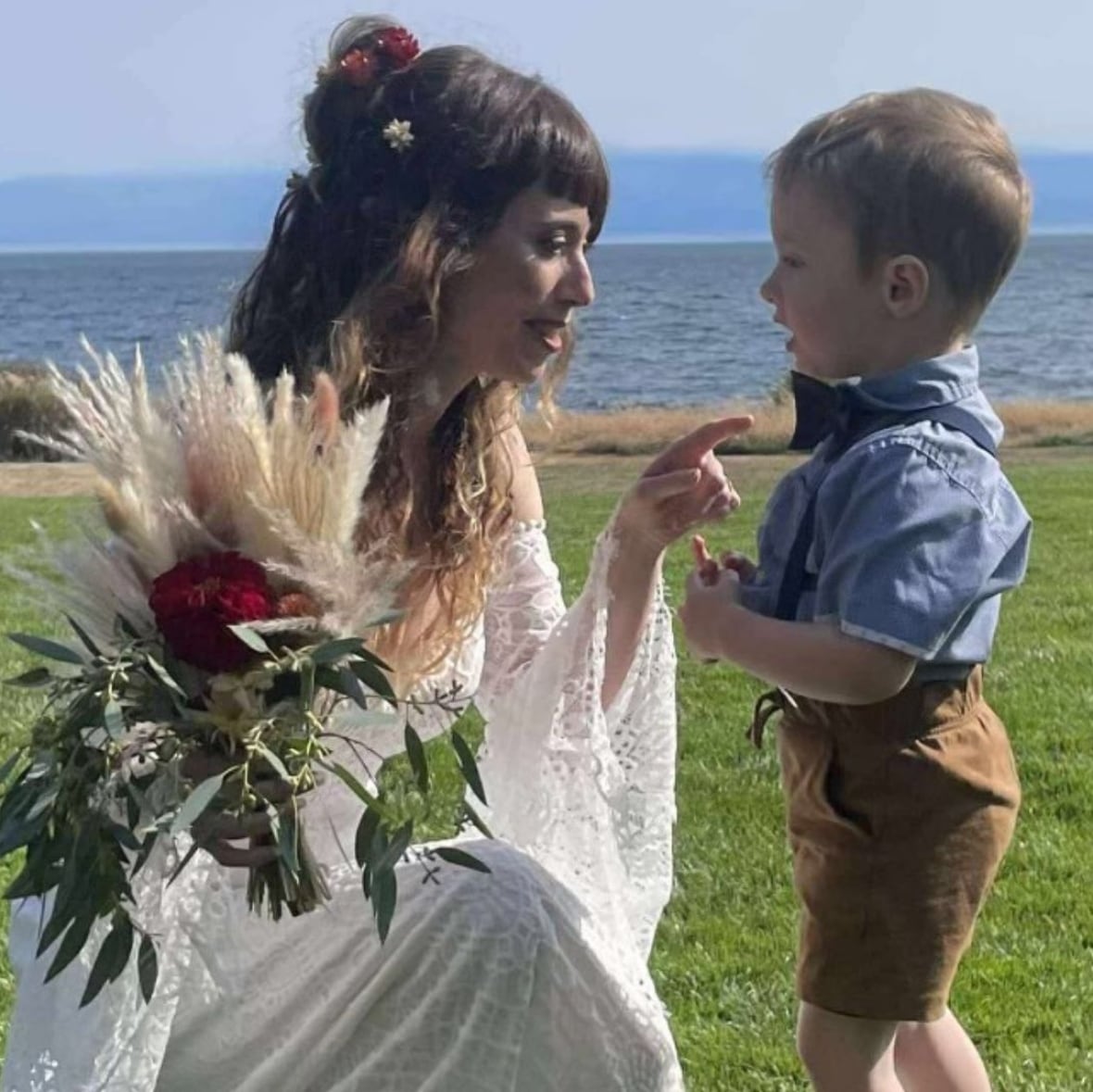A woman in a wedding dress kneels down in a field in front of a young boy who is dressed formally. 