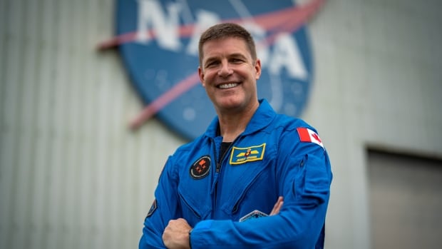 A man in a blue astronaut jumpsuit stands with his arms crossed in front of the blue NASA logo.