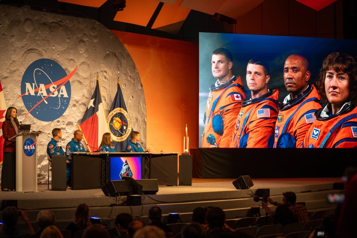 Four astronauts sit in a blue shave on stage and turn to look at a video of themselves in an orange square.