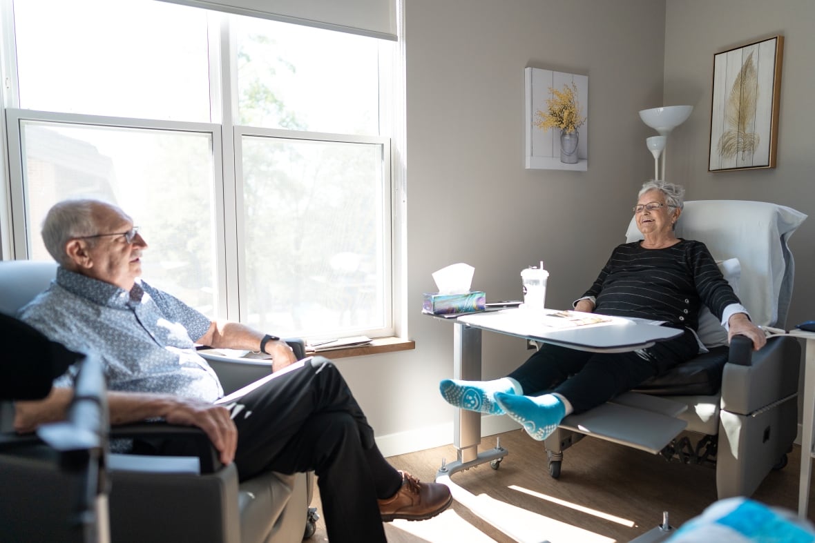 An elderly couple, one in a hospital bed, the other on a sitting chair, have a conversation.