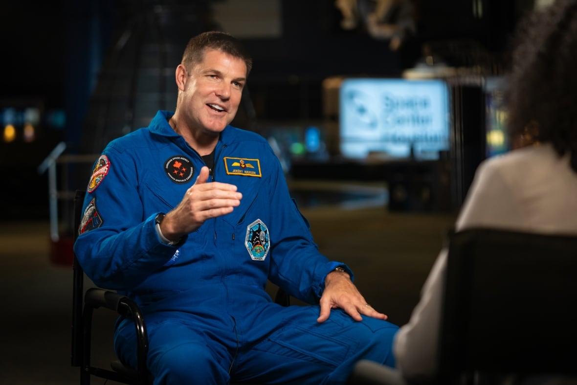 Um homem de macacão azul de astronauta, levanta o braço direito enquanto fala com um repórter.