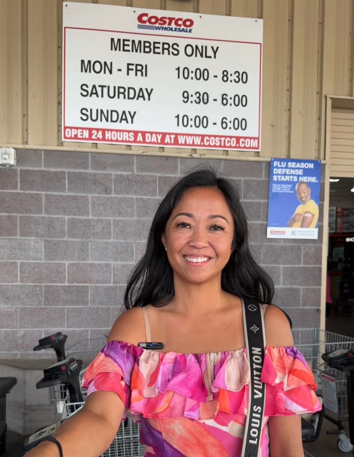 a woman with dark hair and a pink and purple dress stands outside a costco store. behind her the wall of the store and a costco sign are visible, as are some shopping carts