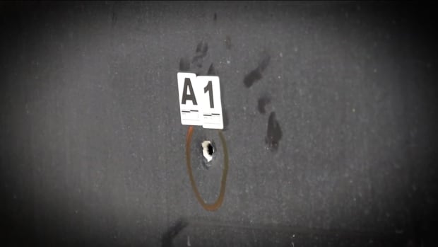 A grey wall with a white bullet hole outlined by an orange circle. There is an evidence marker above the hole with says A1. 