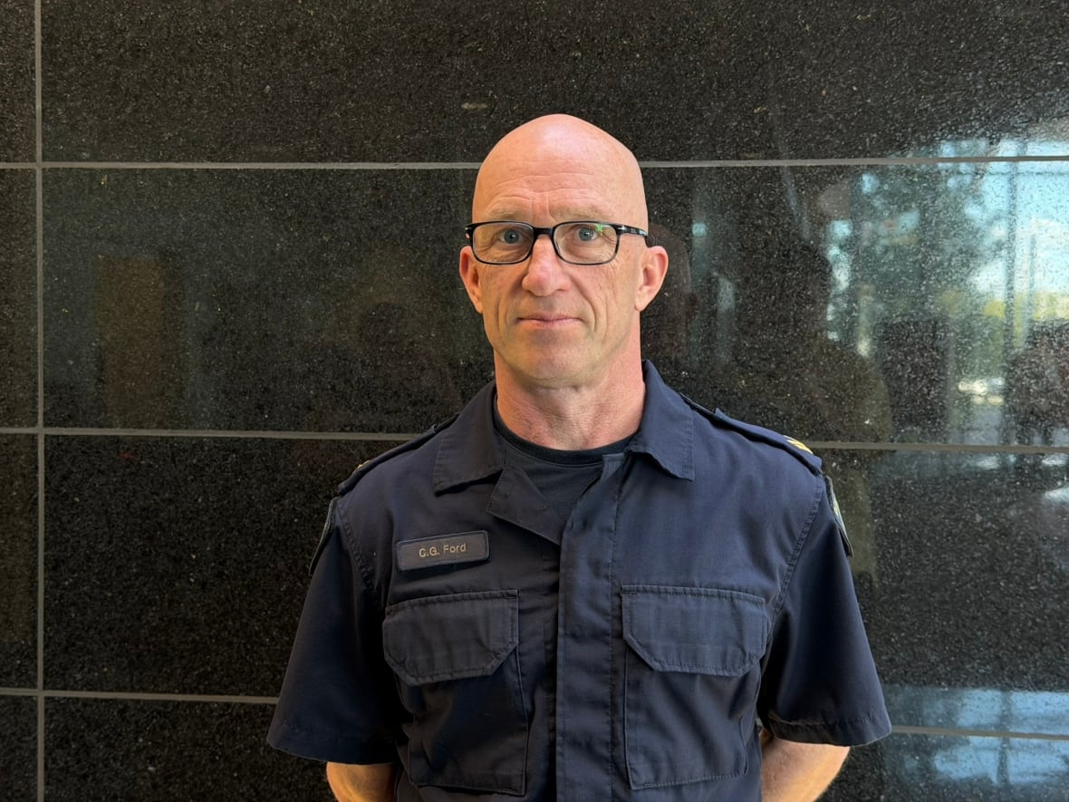 A bald man in a blue RCMP uniform stands in front of a black wall. 