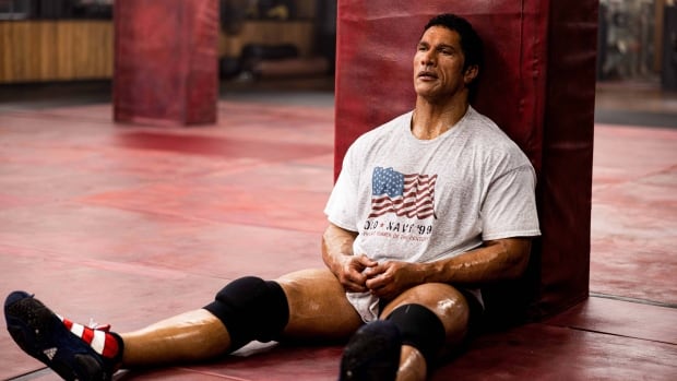 A tired-looking, muscular man leans against a pillar in a wrestling gym.