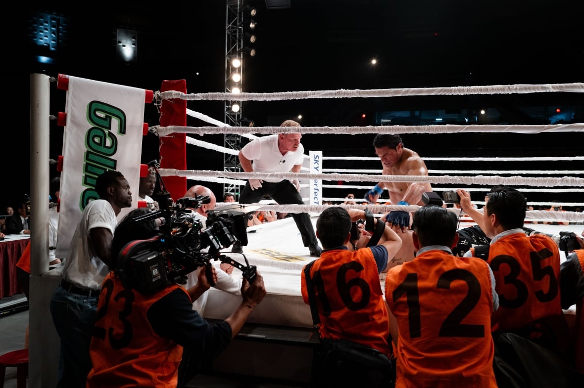 Bir dövüş ringinde bir adam diğerinin üzerinde duruyor. Üstlerinde bir hakem, ringin dışında ise bir grup kameraman var.