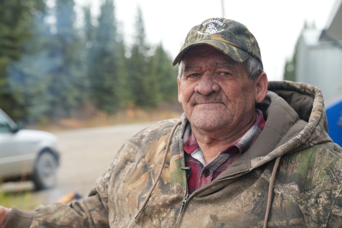 Close-up of a man in camouflage ball cap and jacket, seated with a road behind him and a forest behind the road.  A car on the road enters the frame from the left. 
