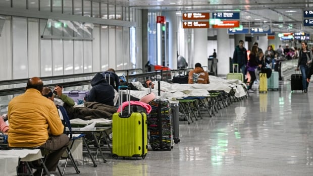 A large indoor space is shown with concrete floors and cots lined up on the side, with people sitting or lying on the cots. Pieces of luggage are shown.