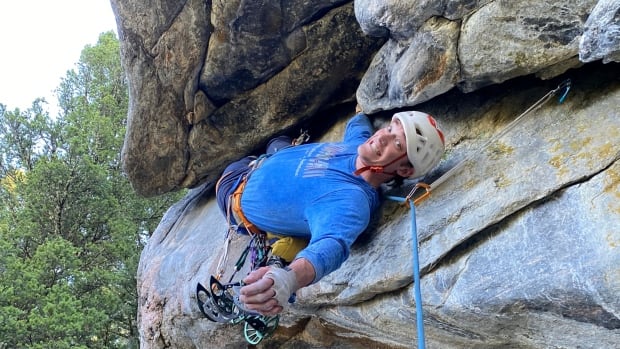 A cleanshaven young man in a helmet smiles as he lays across a rock on a mountain.