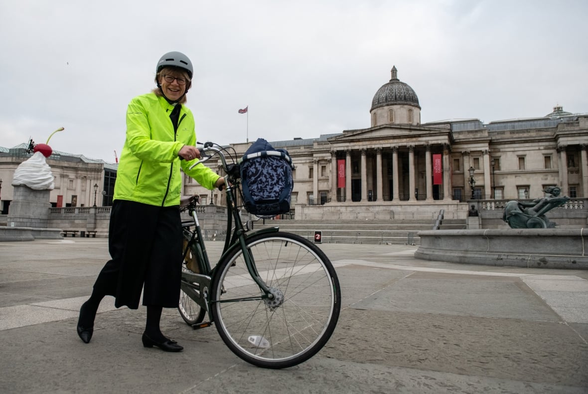 A woman in a yellow reflective jacket and a bicycle helmet is represented standing near a bicycle in a public square.