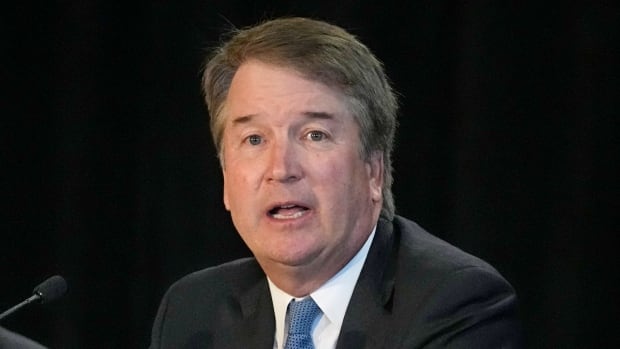 A darkhaired cleanshaven men wearing a suit and tie is shown speaking while seated.