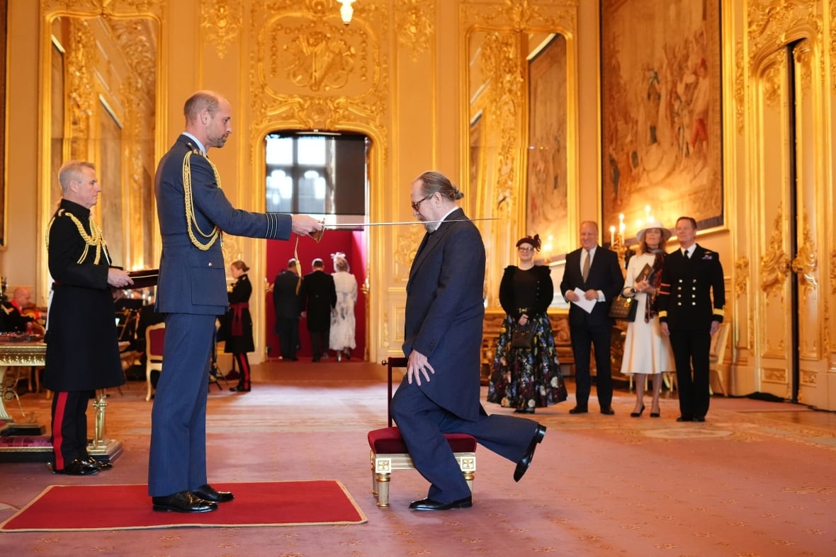 A person kneels on a small stool while another person standing in front of them taps a sword on their left shoulder.