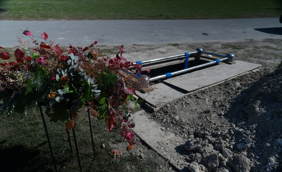 Red and orange flowers sit overtop of an open burial plot, surrounded by plywood boards and a big mound of dirt.