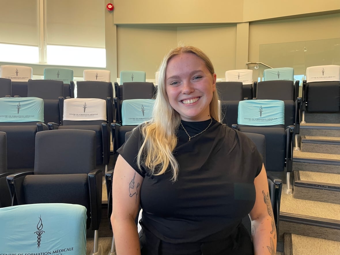A young woman with long blonde hair wearing a black t-shirt smiles, with auditorium seats behind her.