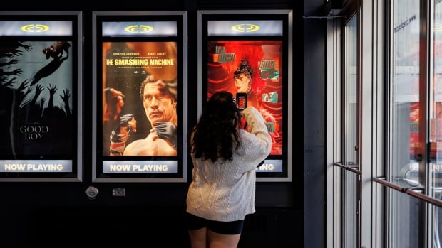 Swifies attend a release party, at Scotiabank theatre, in Toronto, for the launch of Taylor Swift’s The Life of a Showgirl album on October. 3, 2025. a woman in a white sweater takes a picture of a movie poster for taylor swift's official release party of a showgirl theatrical release in a movie theatre