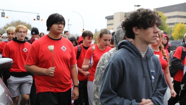 A group of mostly young boys and girls, walk together in dark matching t-shirts in a march, while holding white roses