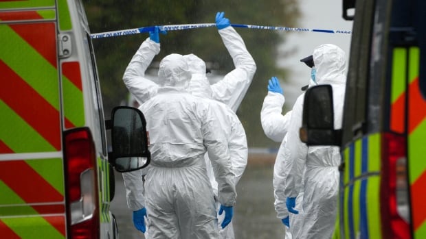 People in forensic suits lift police tape to access an area.