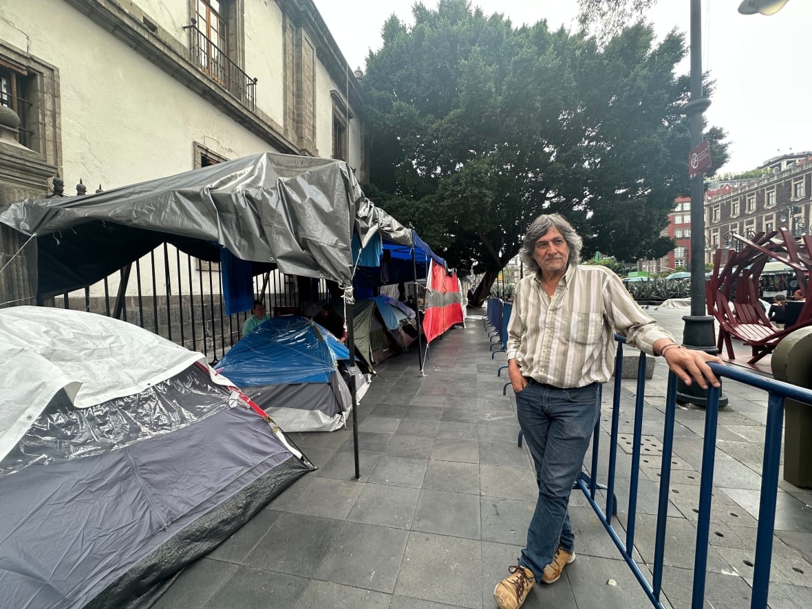 A man stands between a metal fence and a row of small tents. 