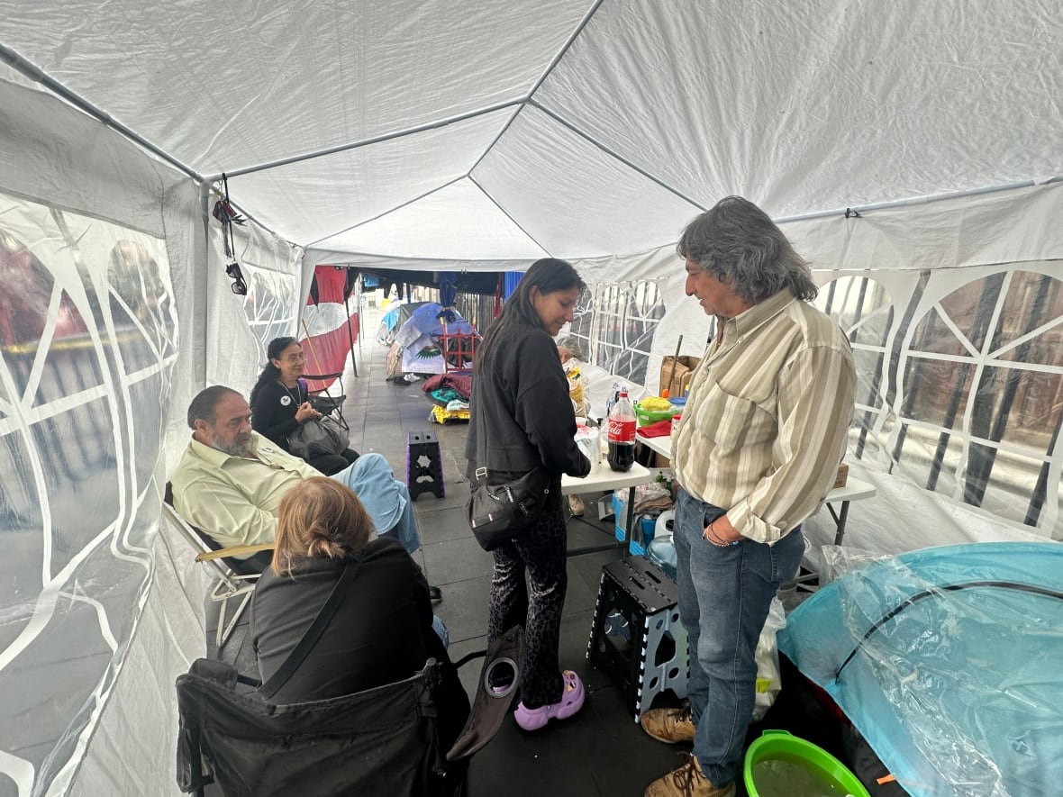A group of people inside a structure similar to a Gazebo. Some people are seated on folding camping chairs talking.