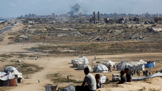 People are seen sitting on a hill overlooking tents and a charred and destroyed city, with smoke still in the air from an explosion.