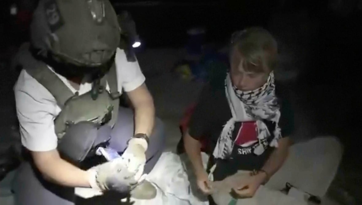 A blonde person carrying a Kffiyeh scarf is at the cross paw next to a soldier wearing a helmet at night.