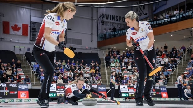 Team Homan skip Rachel Homan, centre, makes a shot as lead Sarah Wilkes, left, and second Emma Miskew sweep during draw six at the PointsBet Invitational in Calgary on Friday. A female curling skip makes a shot as two teammates prepare to sweep while fans watch from the stands.