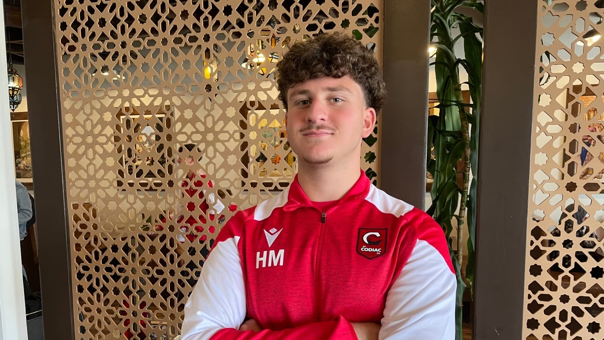 A young man with short curly hair poses with his arms crossed. He's wearing a read and white long sleeved jersey with the initials HM on his chest.