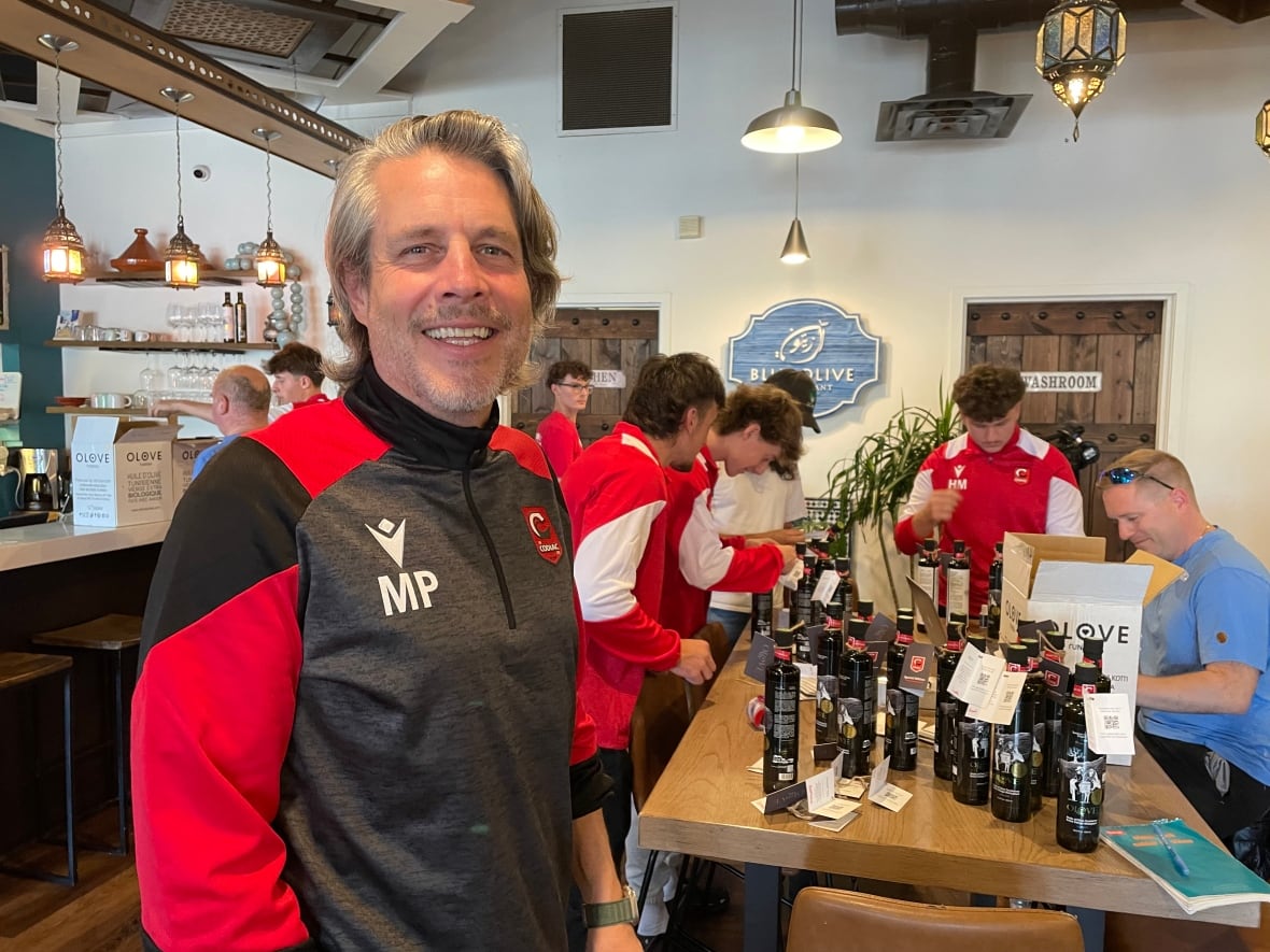 A man with grey hair smiles, standing in front of a table with a group of boys huddled around it. He's wearing a grey and black jersey with the initials MP on his chest.