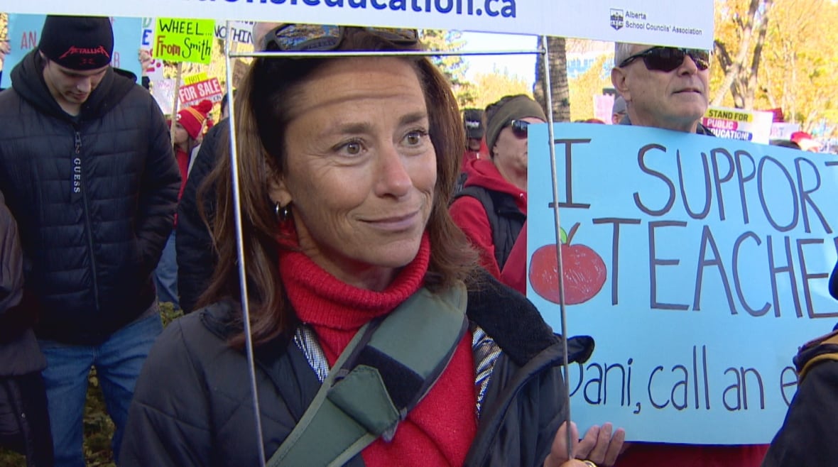 A woman with light skin and shoulder-length brown hair is wearing a red turtleneck under a black jacket. She is standing among a large crowd of people with signs.