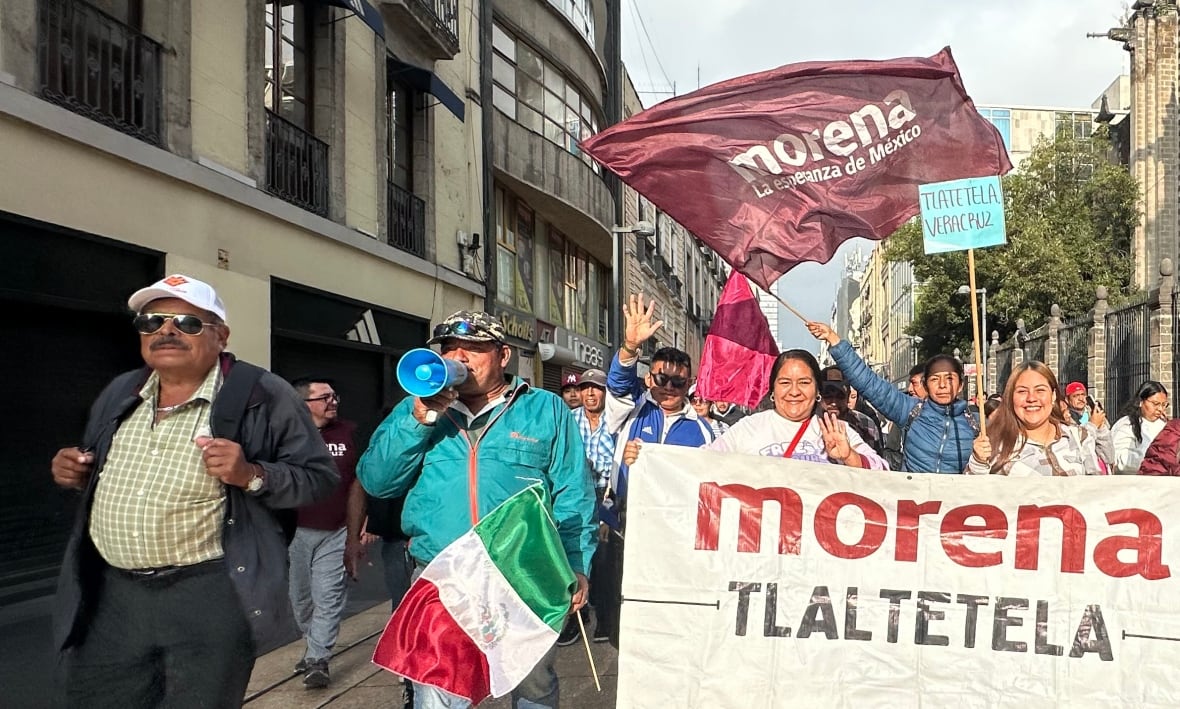 A man with a backpack, a hat and sunglasses walk to the left of a group of people wearing a banner that reads "Morena."