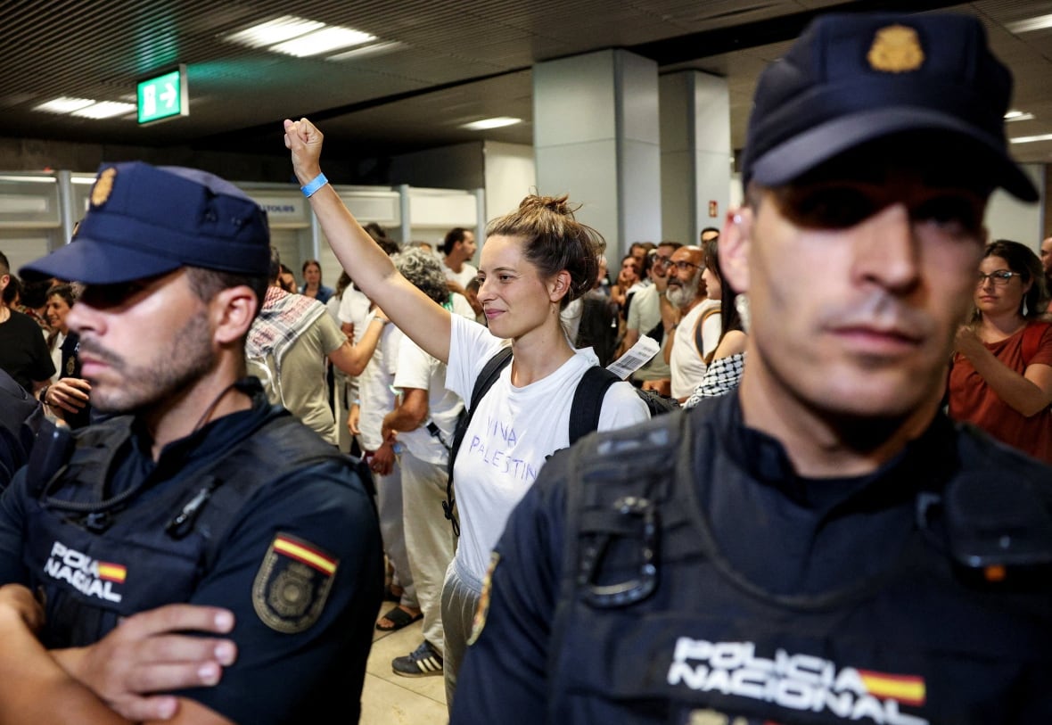 A woman holds a fist while wearing a white T -shirt with FIFA Palestine written handwritten it. Also, the Spanish police hold a crowd.