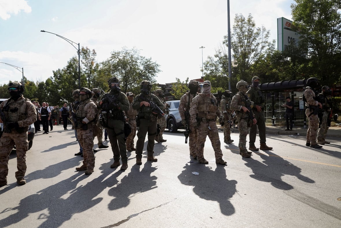 Men masked in military uniforms, holding rifles, stand in the street under the brilliant sun.