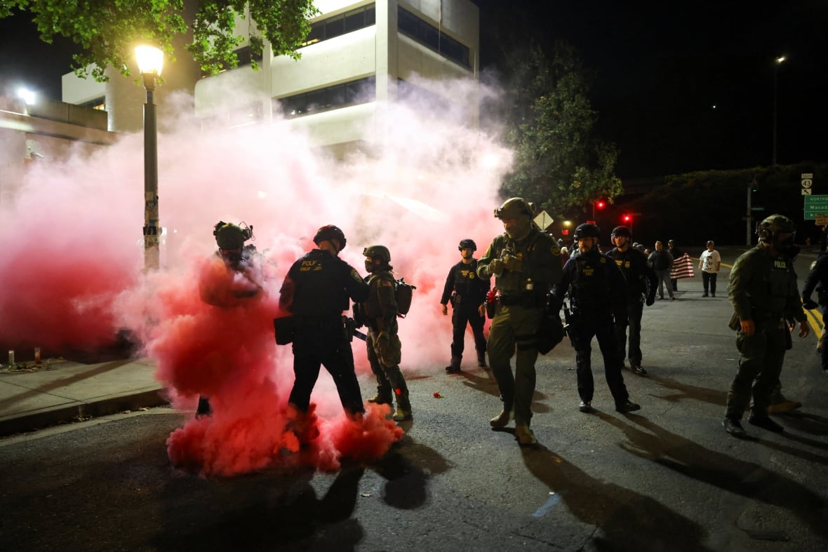 Red smoke revolves around people in the street at night in front of a large white building. Some carry military uniforms.
