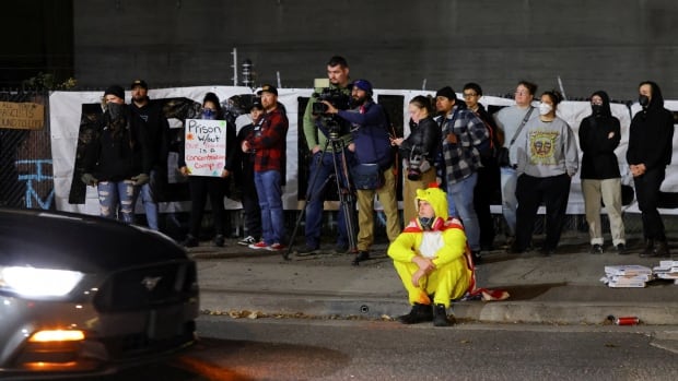 A person wearing a full-body chicken costume sits on the curb of a street in front of a group of protesters with signs and journalists with camera equipment as a car drives past.