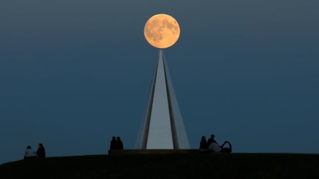 A full moon rising over a pyramid installation at a park, where three groups of people are sitting, just after sunset.