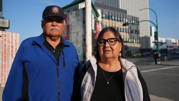 man with a cap on the left, woman with glasses on the right, stand next to a traffic intersection 