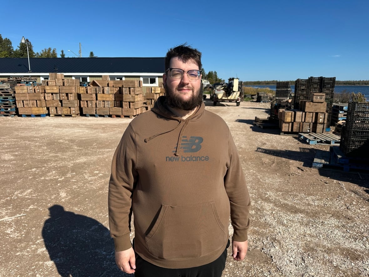 A man in glasses and a brown New Balance hoodie stands outside, in front of palettes holding boxes and a small building.