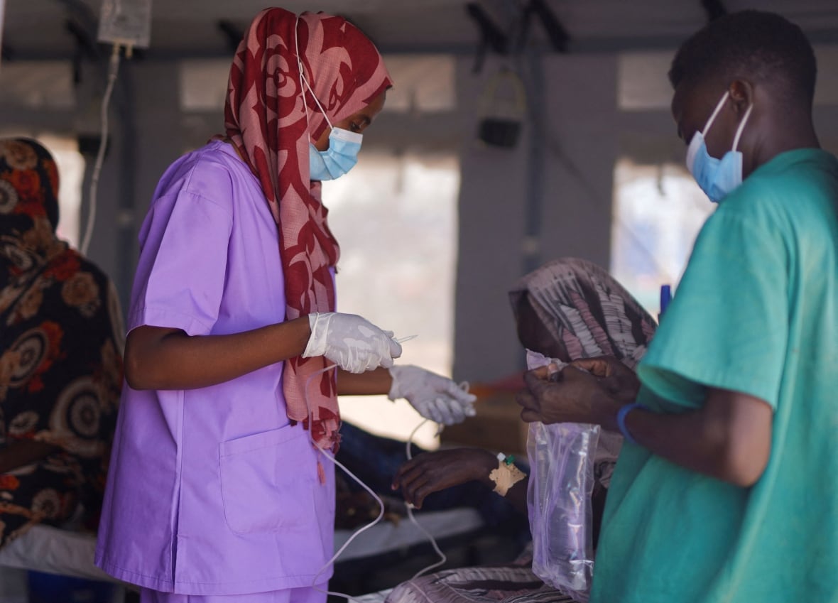 A nurse of males and females, whether wearing scrubs or surgical masks, prepares an IV leak to be managed for a woman sitting in a medical tent. 