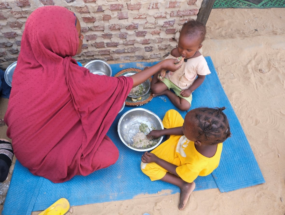 A woman in a red veil sits on a blue blanket feeding on a small handful of food from a silver container while another child sits in the braids next to eating from her own bowl. 