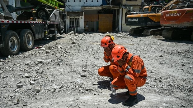 Two men in orange uniforms and helmets kneel on gravel ground and pray, with heavy machinery seen around them.