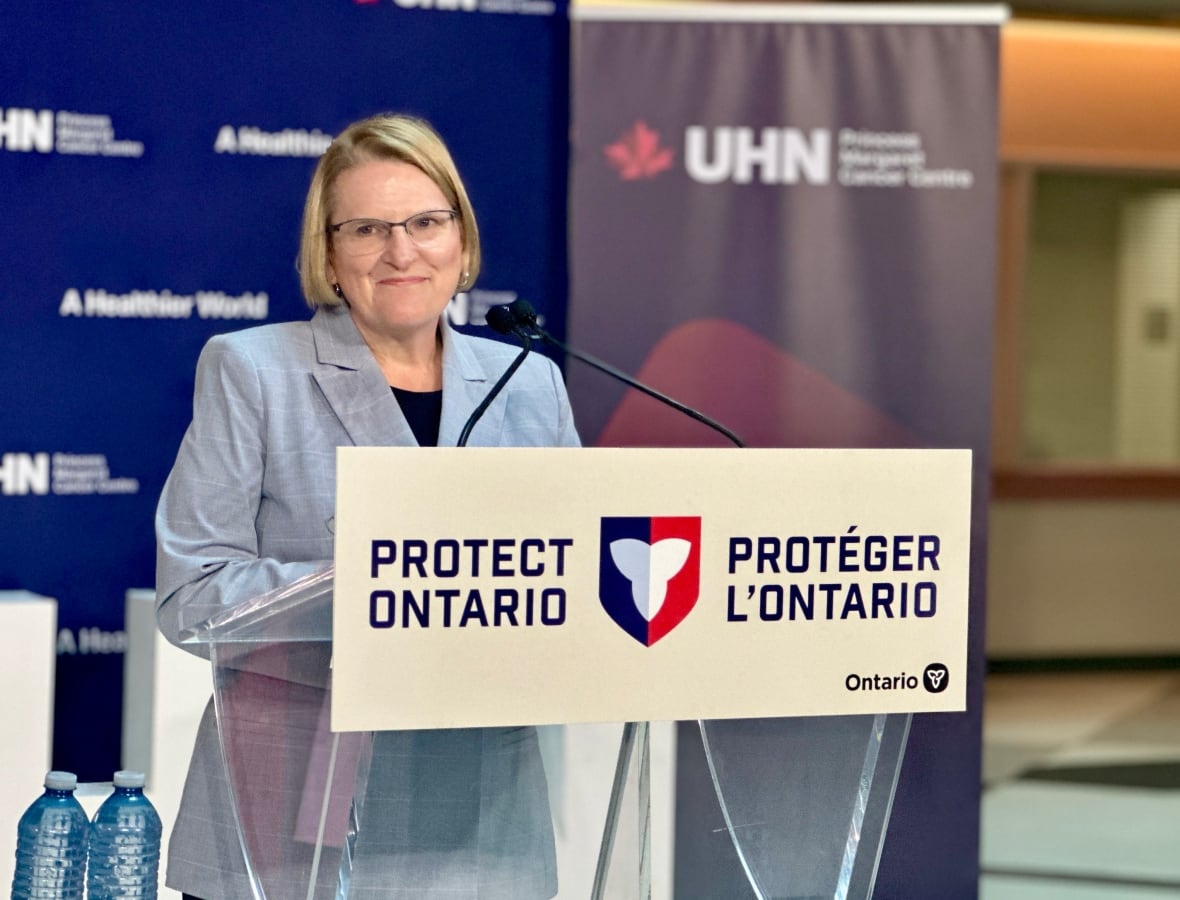 Una mujer con cabello corto y rubio y gafas se encuentra frente a un podio con un cartel que dice Protect Ontario/Proteger L'Ontario.