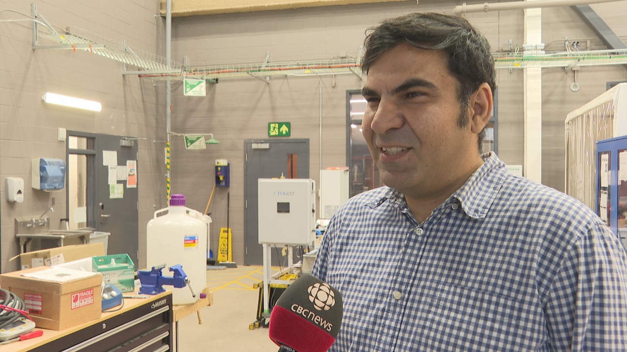 A man in a button-up plaid shirt speaks into a CBC microphone. He is standing in front of a lab.