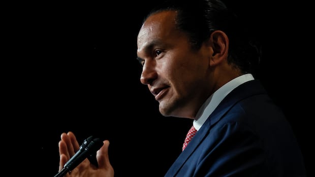 A man in a suit gesticulates with one hand while speaking in front of a mic. On the foreground, there are people seated facing the same direction, but looking at the speaker.