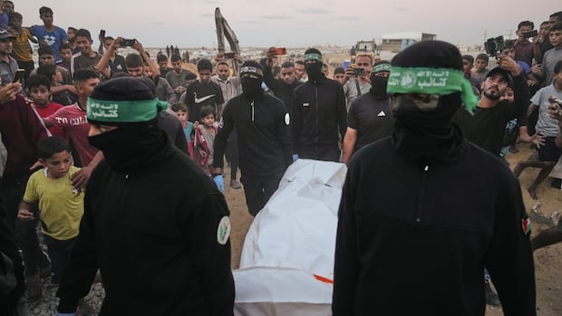 Hamas militants carry a white bag believed to contain a body, after retrieving it from a tunnel during a search for the remains of hostages in Hamad City, Khan Younis, in southern Gaza, Tuesday, Oct. 28, 2025. (AP Photo/Jehad Alshrafi) Militants with face coverings carry a body bag as they are surrounded by people.