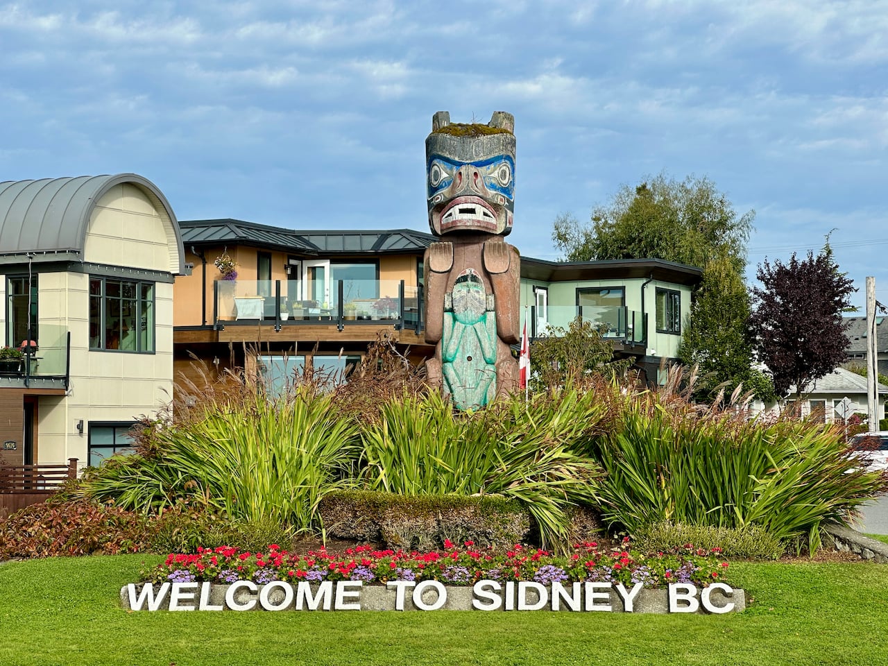 A sign that says "welcome to Sidney," with a totem and buildings in the background. 