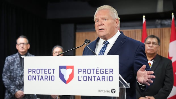 Man stands at podium with microphone in front of a few people and Canadian flags behind