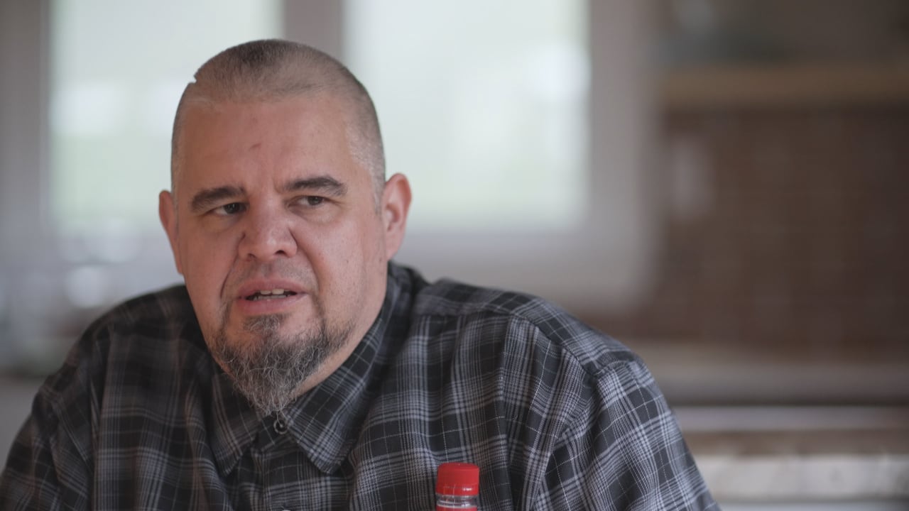 A man in flannel shirt and a beard is talking at a kitchen table