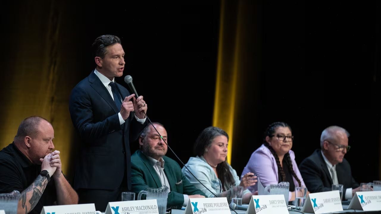 A suited man stands at a microphone, addressing a panel. Five seated individuals listen attentively. The room is dimly lit with a focused spotlight.