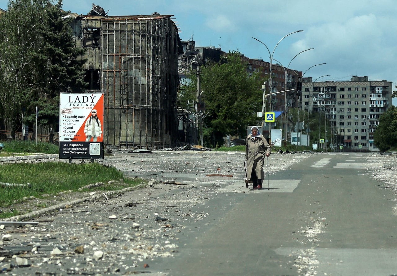 an older woman in a head scarf walks among damaged buildings